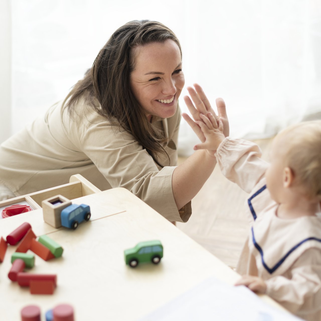 Frau klatscht mit ihrer Hand auf die Hand des Kleinkindes. Dieses sitzt an einem Tisch und spielt mit Bauklötzen und Autos.