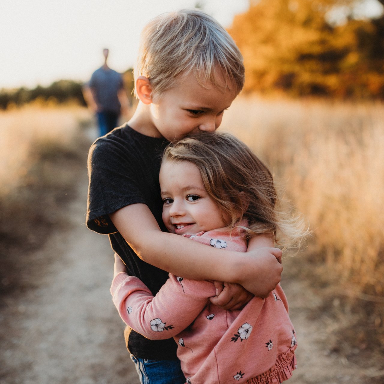 Junge umarmt seine kleine Schwester, im Hintergrund sieht man die Eltern. 